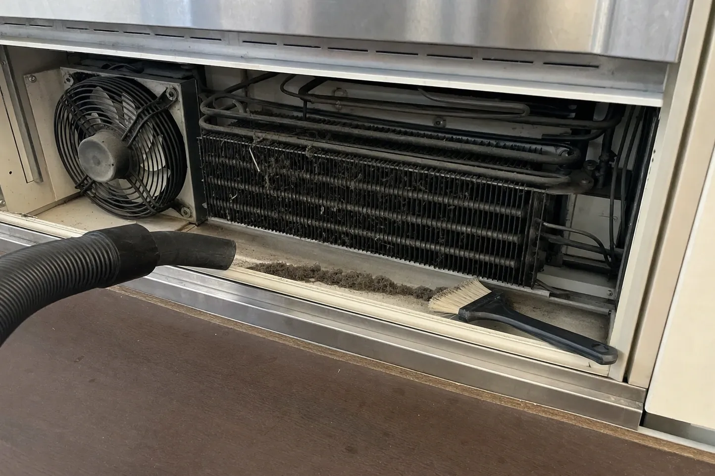 Close-up of a dust-clogged condenser coil and fan behind a removed grille, vacuum nozzle and brush in the gap