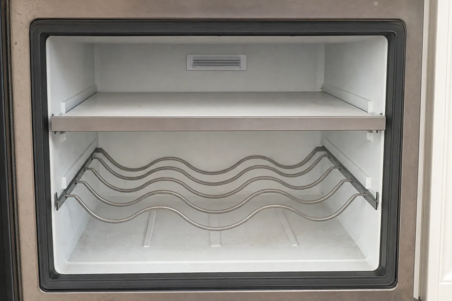 Empty refrigerated compartment behind a thick dark gasket frame, one solid shelf above three wavy wire cradles and a louvred vent panel on the back wall