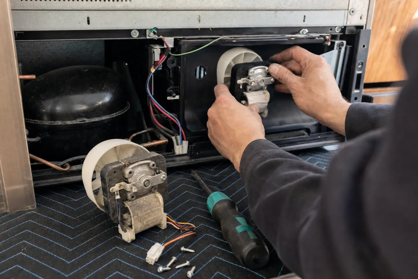 Hands seating a fan motor in its white shroud into an open machine compartment, the dirty original with its leads lying on the floor mat beside four screws